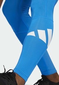 Niebieskie legginsy sportowe wykonane z elastycznego materiału, z białym geometrycznym wzorem na dolnej części nogawki oraz elastycznym pasem w talii. Widoczne czarne sneakersy.