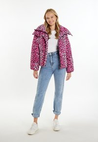 Růžová bunda s leopardím potiskem, zateplená, s vysokým límcem, prošitým texturou, zapínáním na zip a elastickými manžetami, v kombinaci se světle modrými džínami a bílými teniskami.