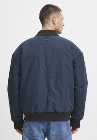 Jūrinės aviacijos bomber tipo striukė su kvadratiniais raštais, įrengtais šonais ir rankovėmis. Savybės: kabliukas nugaroje pakabinimui; medžiaga atrodo lygi ir patvari.