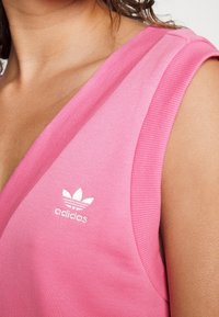 Różowy prążkowany top bez rękawów z dekoltem w kształcie litery V. Posiada białe logo Adidas na klatce piersiowej. Gładka tekstura materiału.