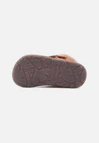 Semelle en caoutchouc marron avec motif texturé, affichant des formes semblables à des feuilles et un logo en relief. Matériau supérieur doux et marron. Design robuste et durable.