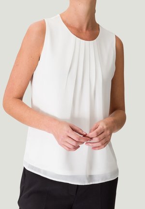 Mouwloze witte blouse met plooien bij de hals, gedragen met zwarte broek, handen zachtjes in elkaar gevouwen voor het lichaam.