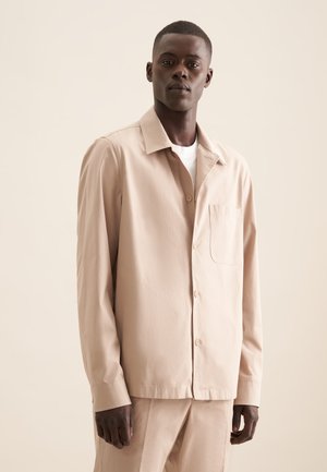 Chemise légère beige clair avec un col, une poche frontale et des boutons. Fabriquée en tissu lisse, elle présente une coupe décontractée et des manches longues.