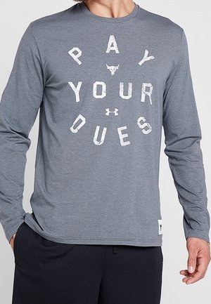 Mann trägt ein graues Langarmshirt mit "PAY YOUR DUES" in weißen Buchstaben und kleinen Logos auf Brust und Saum.