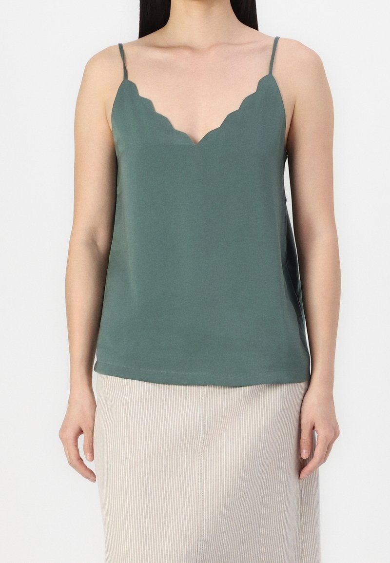 Femme portant un haut caraco vert à col en V festonné avec de fines bretelles et une jupe côtelée beige clair.