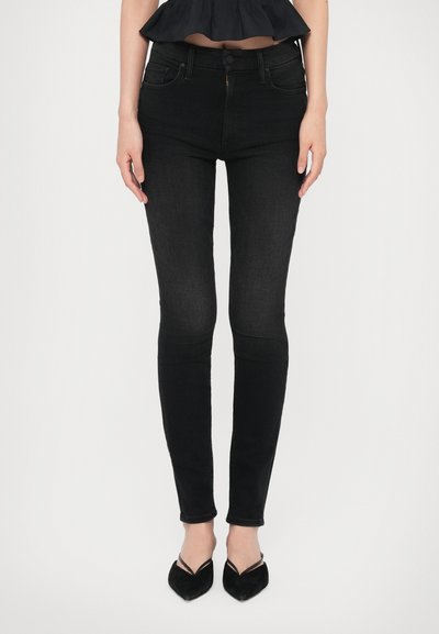 Femme portant un jean skinny noir taille haute avec des poches avant et des chaussures à talons aiguilles noires, se tenant contre un fond blanc.