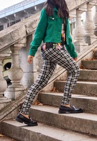Grüne strukturierte Jacke, Blumenhemd, schwarz-weiße karierten Hosen und schwarze Plateauschuhe, die steinerne Treppen mit dekorativem Geländer hinaufgehen.
