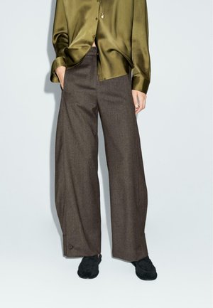 Personne portant un pantalon large marron foncé, une blouse en satin vert olive, des chaussures noires et un collier pendentif, debout devant un fond uni.