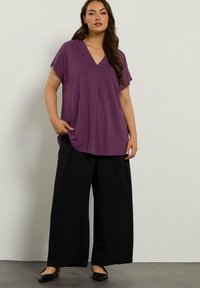 Lila V-Ausschnitt-Oberteil mit kurzen Ärmeln, kombiniert mit schwarzen weiten Hosen. Glattes Material mit lässiger, lockerer Passform. Schwarze Ballerinas ergänzen das Outfit.