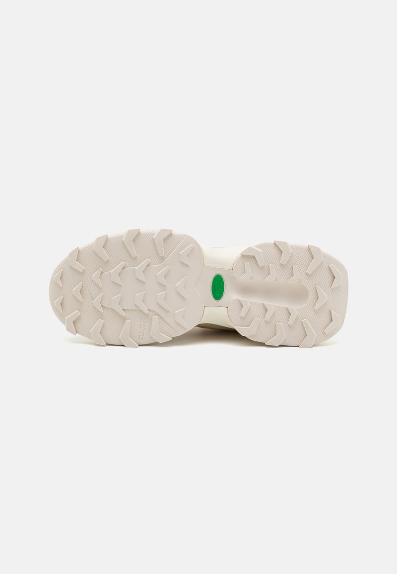 Semelle de sneaker couleur crème avec un caoutchouc texturé présentant des motifs de bande de roulement proéminents et un accent de logo vert. Conçue pour la traction et la durabilité.