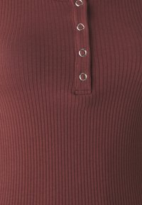 Burgundská rebrovaná tielko s henley výstrihom, s piatimi kovovými gombíkmi na prednej strane. Mäkkučká, textúrovaná tkanina.
