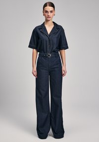 Dungaree in denim di colore blu scuro con maniche corte, collo a camicia, chiusura con bottoni e vita con cintura. Presenta un design a gamba larga.