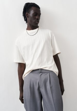 Homme à la peau foncée avec des dreadlocks portant un T-shirt oversize blanc à fines rayures, un pantalon plissé gris et un collier chaîne argenté, debout contre un mur blanc.