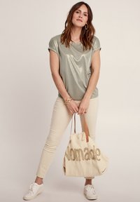 Maglietta lucida verde oliva con maniche arrotolate, abbinata a pantaloni aderenti beige chiaro, sneakers bianche e una grande borsa crema con la scritta "Nomade" in lettering dorato.