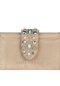 Cartera de ante beige con un cierre decorativo que presenta varios remaches y una gema azul. Texto "ALMA EN PENA" grabado en la parte delantera.