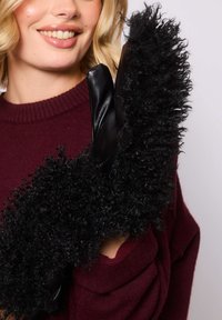 Una donna che indossa un maglione rosso scuro tiene in alto un paio di guanti di pelle nera con un lungo bordo di pelliccia riccia, sorridendo leggermente.