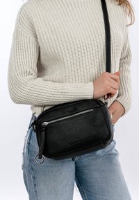 Osoba ubrana w kremowy sweter na drutach i niebieskie dżinsy, trzymająca czarną skórzaną torbę na ramię z srebrnymi zamkami błyskawicznymi.