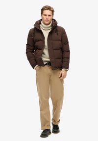 Chaqueta de plumas marrón con capucha, cierre de cremallera y cordón. Lleva puesta una camiseta de cuello alto crema. Pantalones beige y zapatos negros completan el conjunto.