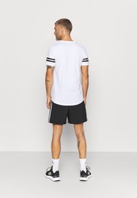 T-shirt bianco a maniche corte con strisce nere sulle maniche, abbinato a pantaloni corti neri con strisce bianche ai lati e scarpe da ginnastica nere.