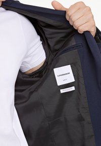 Chaqueta azul marino con corte ajustado, con un forro interior negro, y etiquetas de especificaciones de marca y material visibles en el interior.