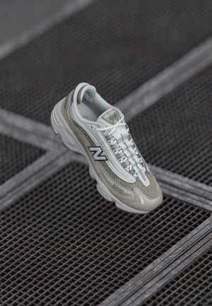 New Balance-sneaker med en beige og hvit mesh-overdel, strømlinjeformet fasong, synlige lisser og subtile merkevare detaljer, plassert mot et rutenettbakgrunn.