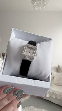 Woman's hand with metallic silver nails holding a Guess watch with a square, silver, rhinestone-encrusted face and black strap in a white box.