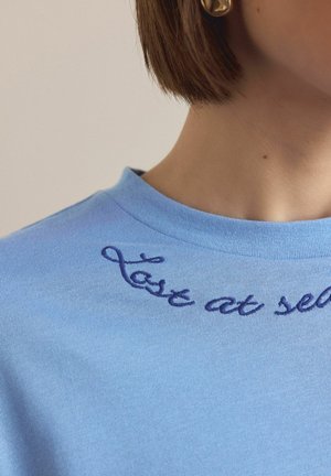 Lyseblå bomulds-T-shirt med rund hals og et buet, marineblåt broderet tekstdesign, der lyder "last at self."