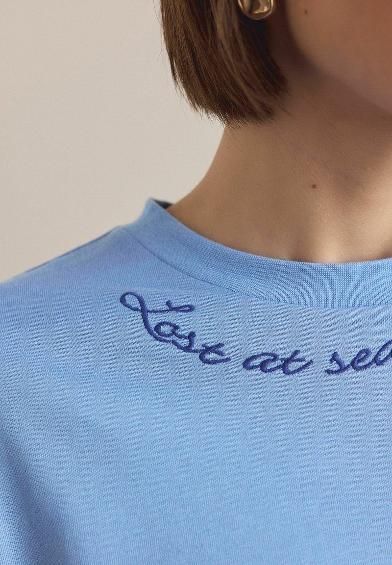 Svetlo modra bombažna majica z okroglim izrezom in ukrivljenim vezenim napisom v temno modri barvi, ki se glasi "last at self."