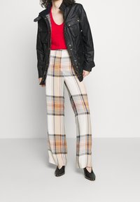 Giacca in ecopelle nera con due tasche sul petto, top rosso a scollo a V e pantaloni a gamba larga a quadri con motivi beige, marroni e navy.