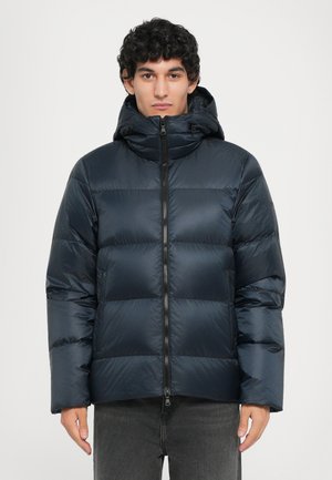 Navy puffer jacket with horizontal quilted design, zip closure, adjustable hood, and smooth, shiny texture. Model wearing gray pants.