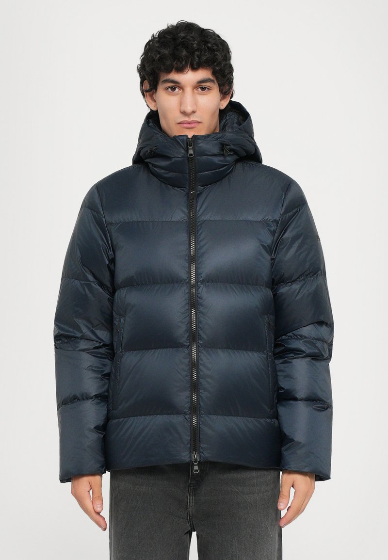 Marineblauer Pufferjacke mit horizontal gestepptem Design, Reißverschluss, verstellbarer Kapuze und glatter, glänzender Textur. Model trägt graue Hose.