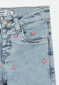 Jeans in denim azzurro chiaro con piccoli ricami a forma di cuore rosa sparsi vicino alla tasca anteriore e alla vita.