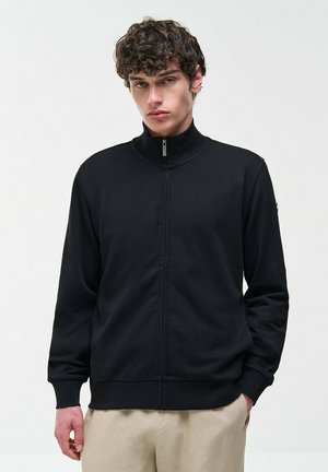Giovane uomo con capelli ricci, che indossa una giacca nera con cerniera e pantaloni beige, in piedi davanti a uno sfondo chiaro e uniforme.