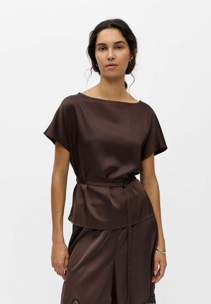 Femme portant un haut court en satin marron foncé à manches courtes et un pantalon assorti avec une ceinture, debout devant un fond clair uni.