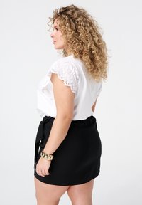 Haut blanc à manches en œillets festonnés, associé à une jupe noire texturée avec une taille à nouer. Bracelet doré au poignet.