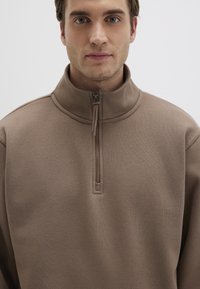 Brown zip-up sweatshirt with a high collar, featuring a smooth texture and a half-zip front closure. Simple, minimalist design.