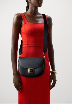 Black leather crossbody bag with a rounded shape, glossy texture, gold hardware, and a brand nameplate. Worn with a fitted red dress.