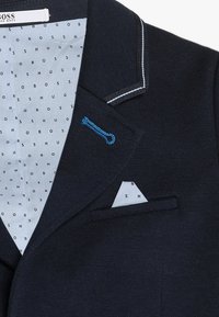 Granatowy blezer z rozciętym klapą, białym przeszyciem i niebieskim akcentem. Wnętrze posiada wzorzystą podszewkę z powtarzającym się napisem „BOSS”. W zestawie poszetka.