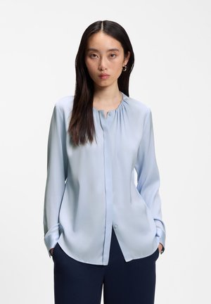 Femme aux longs cheveux foncés portant un chemisier bleu clair et un pantalon foncé, debout les mains dans les poches devant un fond uni.