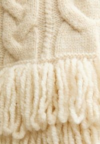 Miękka kremowa dzianina z wzorami warkoczy oraz grubymi, pętlami frędzlami na krawędzi.