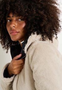 Chaqueta de piel sintética crema con interior negro, capucha y herrajes plateados; presenta una superficie texturizada y un cierre lateral con cremallera.