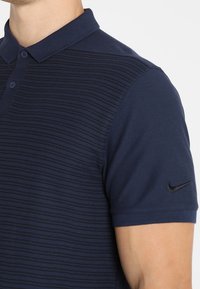 Homme portant un polo bleu marine à rayures horizontales texturées et un logo Nike noir sur la manche gauche.