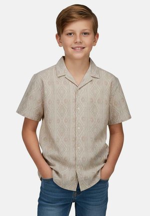 Jongen met kort lichtbruin haar die lacht, draagt een beige overhemd met korte mouwen en patroon en een blauwe spijkerbroek, met handen in de zakken.