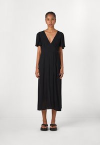 Robe maxi noire avec un décolleté en V profond, des manches courtes et volantes, et une coupe décontractée. Tissu léger avec une texture subtile, associée à des sandales noires.