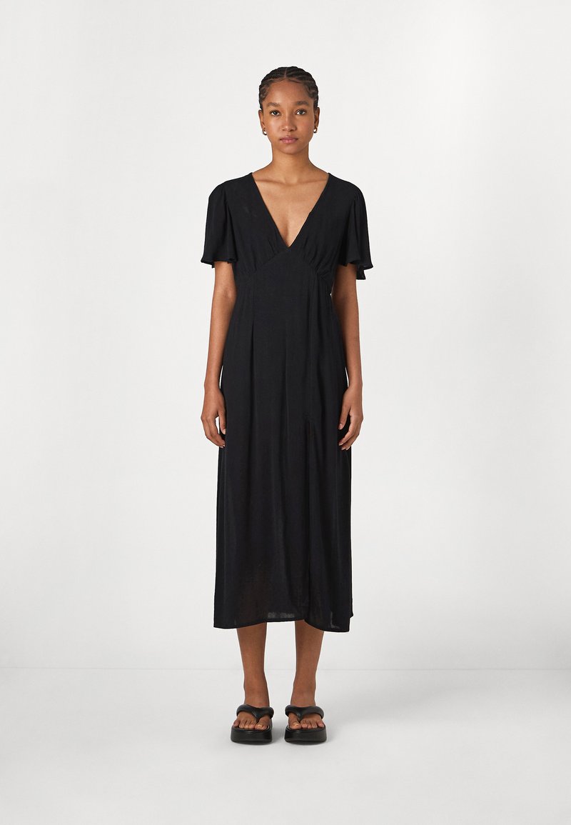 Robe maxi noire avec un décolleté en V profond, des manches courtes et volantes, et une coupe décontractée. Tissu léger avec une texture subtile, associée à des sandales noires.