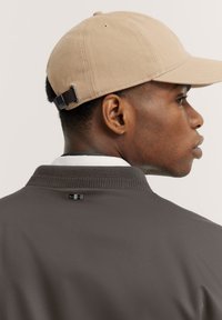 Braune Bomberjacke mit geripptem Kragen, ausgestattet mit einem schwarzen Logo-Tag auf der Rückseite. Beigefarbene Mütze mit verstellbarem Riemen auf der Rückseite.