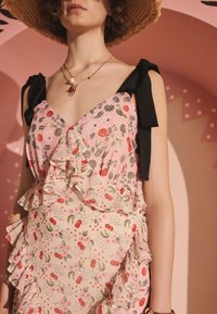 Kvinna som bär en rosa klänning med blommönster och körsbär, svarta axelband, flerskiktade guldkedjor och en vävd halmhatt.