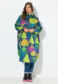 Vrouw staat in een kleurrijke marineblauwe regenjas met neon boompatronen, blauwe opgerolde jeans en zwarte laarzen met roze zolen.