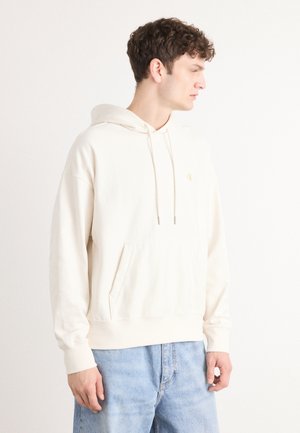 Crème hoodie van zacht materiaal met een voorzak, trekkoordcapuchon en geborduurd logo in het goud. Gepaard met blauwe jeans.