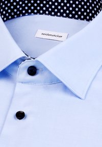 Camicia di colore azzurro chiaro con colletto strutturato, caratterizzata da bottoni neri e un motivo a pois navy all'interno del colletto. Etichetta visibile.